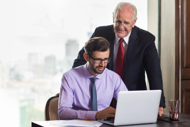 senior business man looking at how another younger man works 1262 778